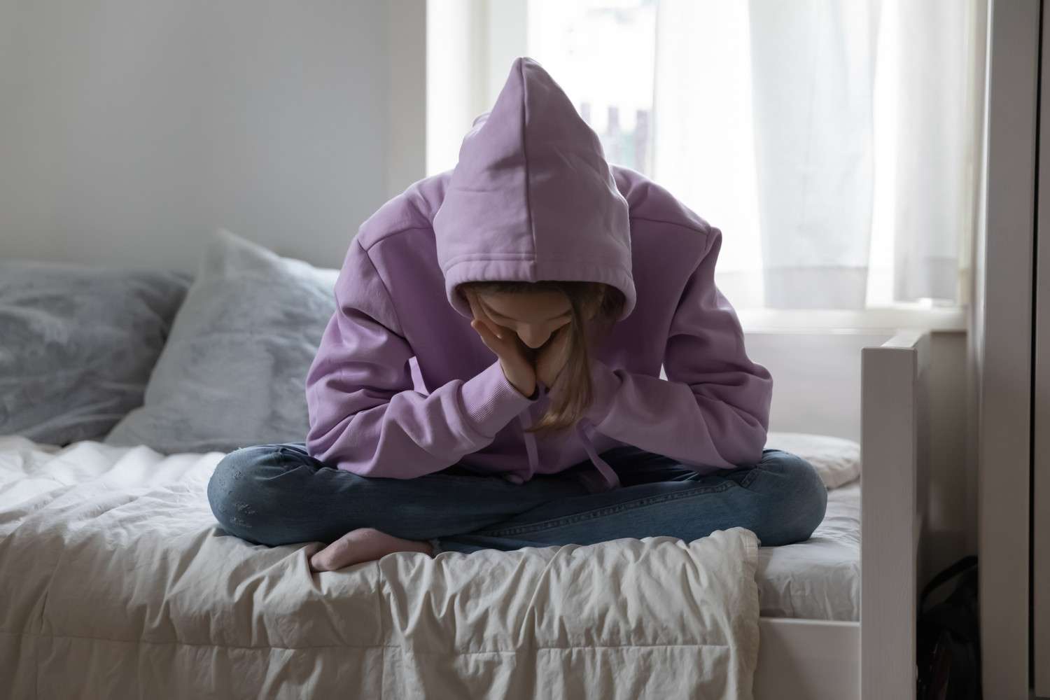 Unhappy teenage girl sitting alone on bed, wearing a sweatshirt and looking depressed.